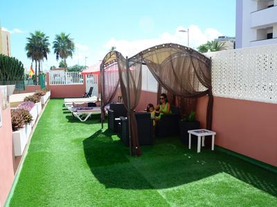 Hotelová terasa se zeleným umělým travnatým kobercem, lehátky a zastřešenými posezeními pod modrou oblohou