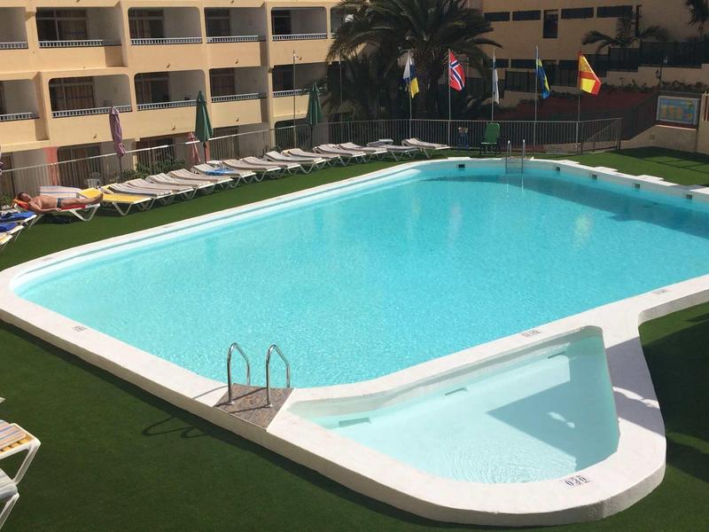 Obdelníkový bazén s vedlejším dětským bazénem, obklopený lehátky a trávníkem.