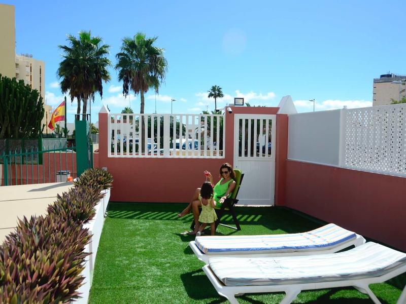 Dva lidé sedící na terase s lehátky a umělým trávníkem pod modrou oblohou.