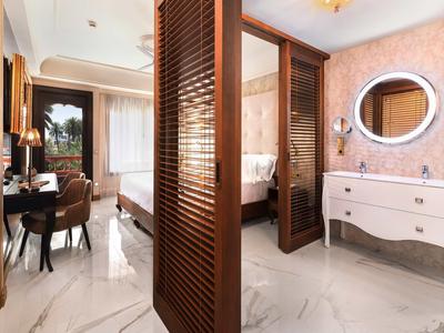 Elegancki pokój hotelowy z łóżkiem, biurkiem oraz łazienką z białymi meblami i owalnym lustrem.