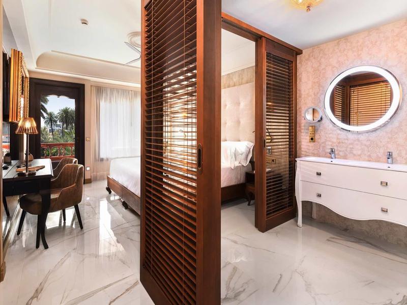 Elegancki pokój hotelowy z łóżkiem, biurkiem oraz łazienką z białymi meblami i owalnym lustrem.