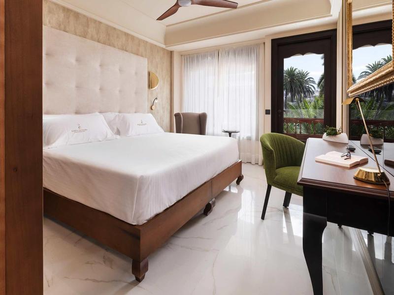 Elegancki pokój hotelowy z podwójnym łóżkiem, zielonym fotelem i drewnianym biurkiem przy oknie.