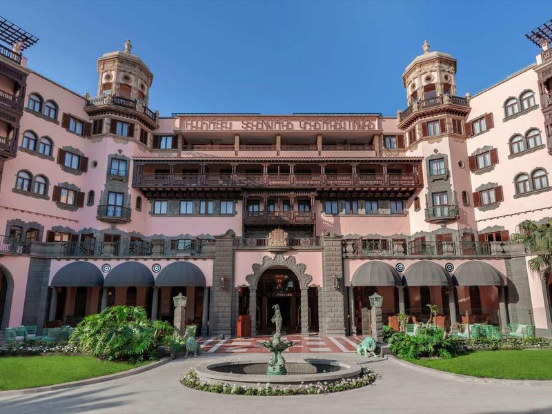 Historyczny hotel z dwiema wieżami, centralną fontanną i ogrodem na tle jasnoniebieskiego nieba.