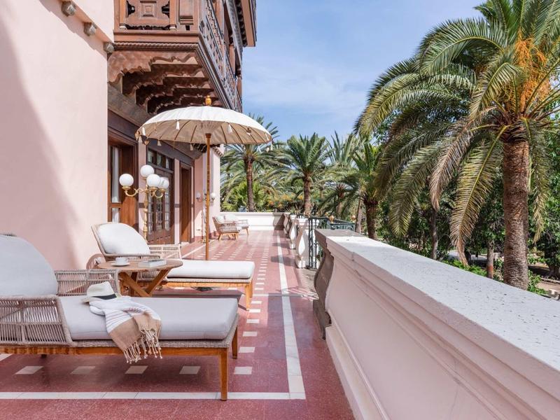 Balkon hotelowy z leżakami, parasolem i palmami pod błękitnym niebem.