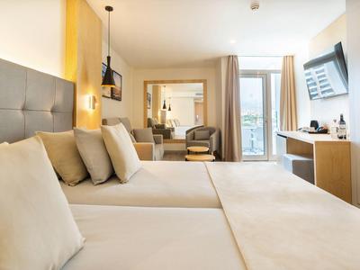 Lichte hotelkamer met twee bedden, fauteuils, tv en balkon met uitzicht op buiten.