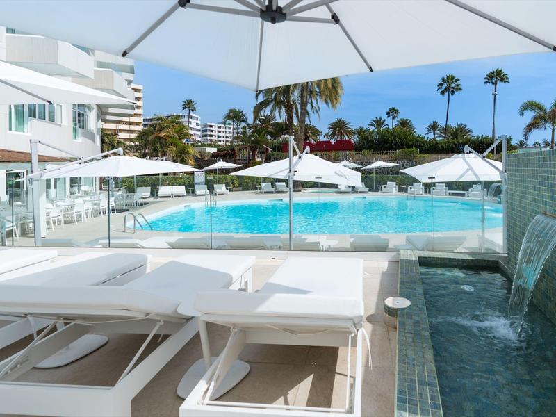 Moderne hotelzwembad met witte parasols, ligbedden en waterval aan de rand.