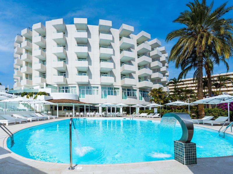 Rond hotel met witte gevel en blauw zwembad vooraan onder blauwe lucht.