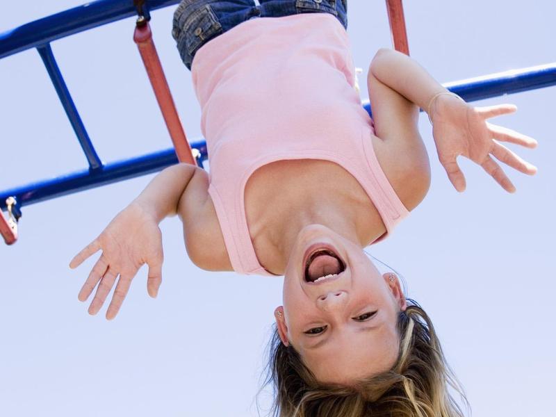 Een blij kind hangt ondersteboven aan een blauwe klimrek buiten met een blauwe lucht.