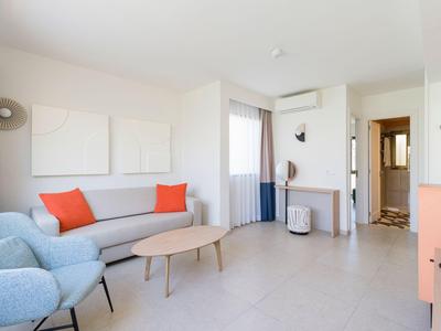 Bright hotel room with sofa, blue armchairs, wooden table, and desk by window.