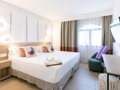 Bright hotel room with double bed, nightstands, seating area, and large window with curtains.