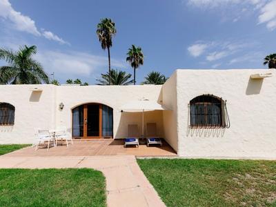 A white, flat-roofed vacation house with wooden doors and surrounding greenery under a blue sky.