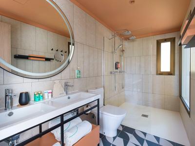 Bright bathroom with double sinks, large round mirror, toilet, and walk-in shower.