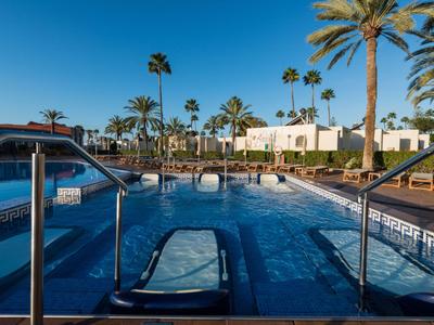 Modern pool with loungers, palm trees, and buildings under clear sky in a vacation setting.
