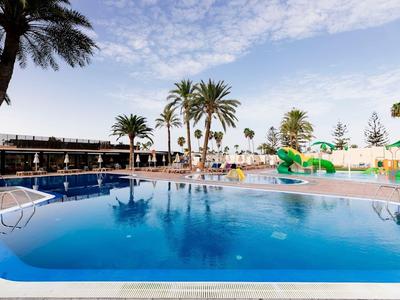 Large outdoor pool with palm trees and water slides under blue sky at a hotel resort.