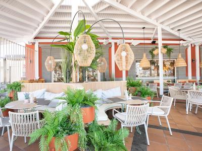 Bright restaurant area with white chairs, plants, and decorative lamps under a wooden roof.