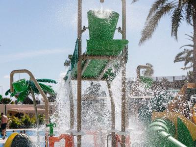 Water slide with large green slide and splashing water, surrounded by palm trees.