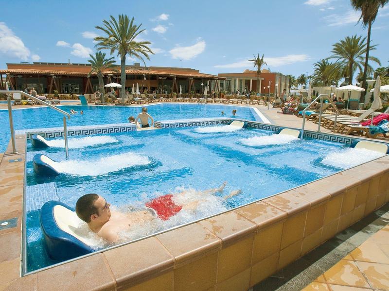 Person relaxing in a hot tub beside a large pool with loungers and palm trees at the hotel resort.