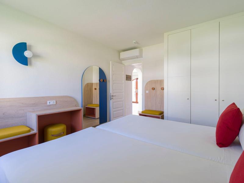 Modern hotel room with large bed, red pillows, wardrobe, and yellow seating.