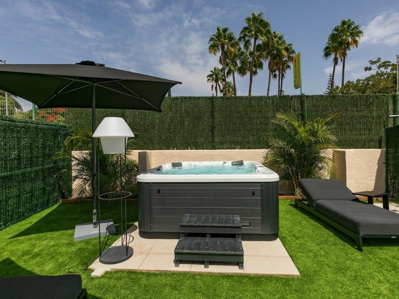 Modern hot tub on a terrace with lounge chair, sunshade, and green hedge.