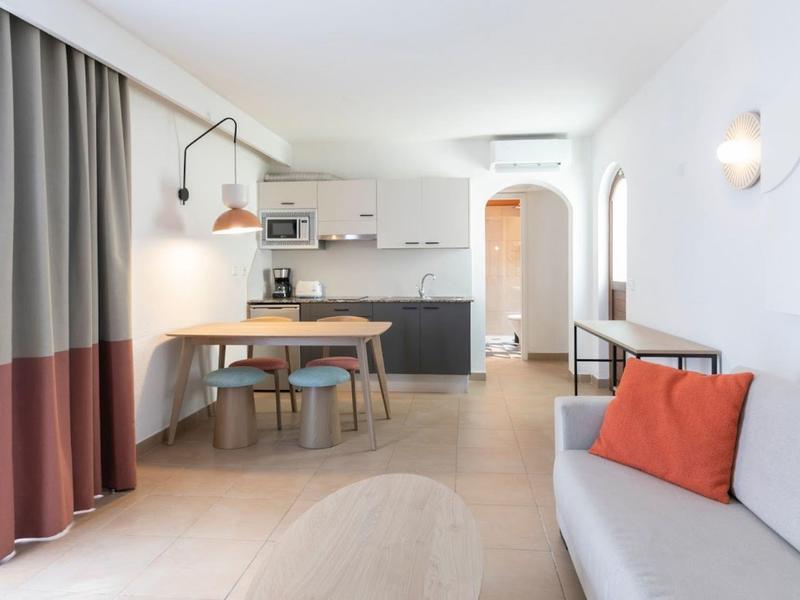 Bright hotel room with modern kitchen, dining table, sofa, and curtains in muted colors.