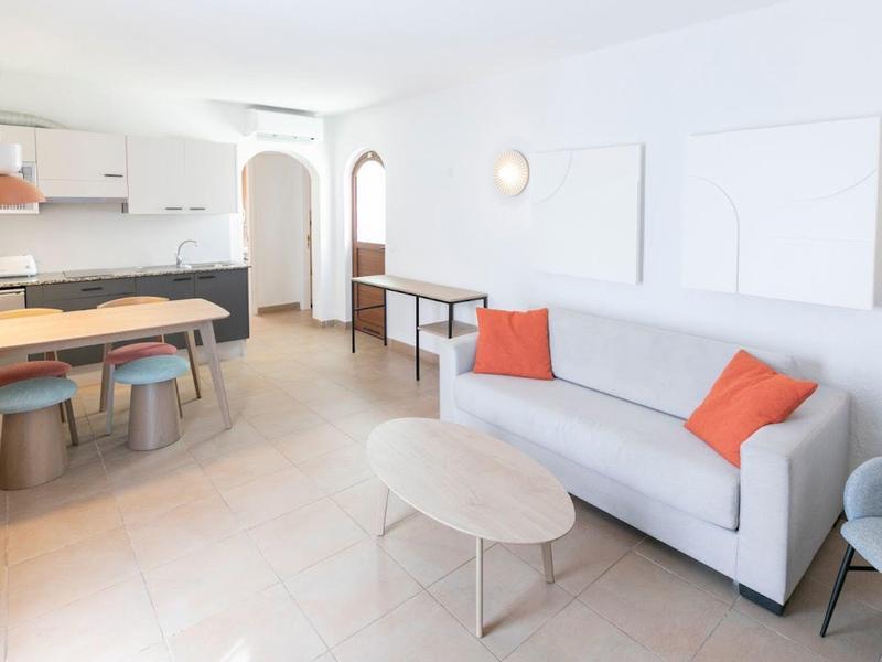Bright living room with gray sofa, orange cushions, dining table, and modern kitchen in the background.