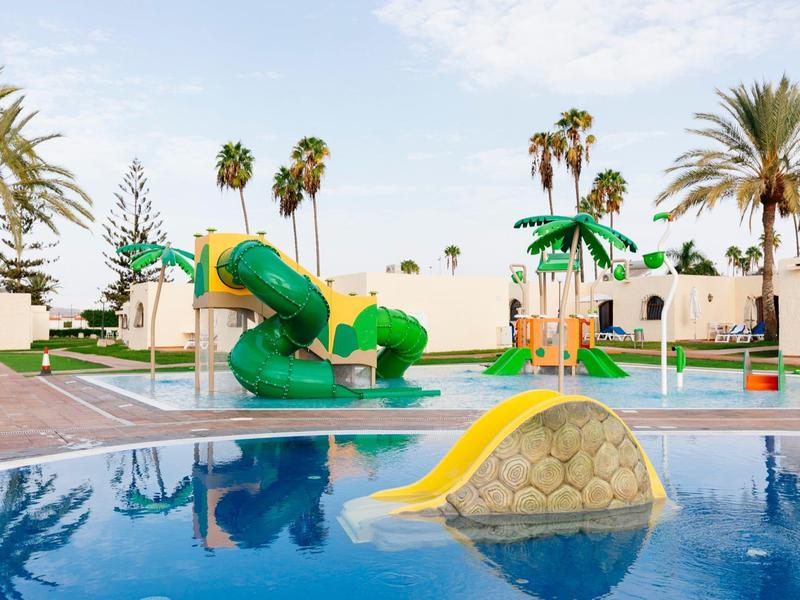 Children's pool with green water slide and yellow climbing rock under palm trees at a holiday resort.