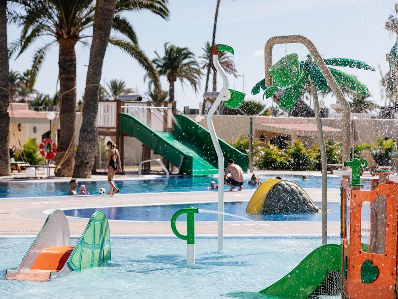 Children's play area with water slides and palm trees in an outdoor hotel pool.