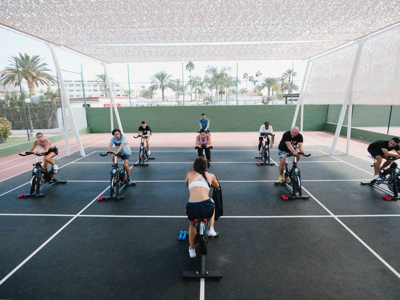 Group of people exercising on indoor spinning bikes under a roof with palm trees in the background.