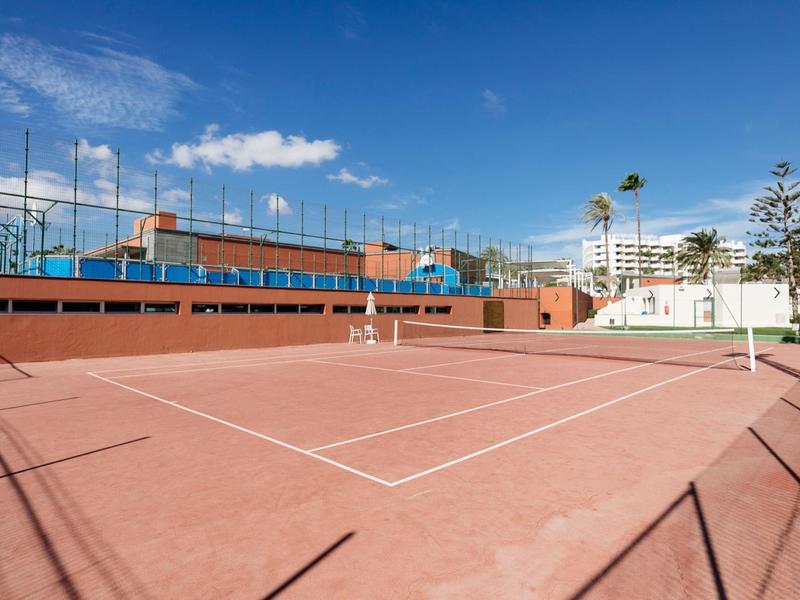 Red clay tennis court with fences and buildings in the background under blue sky.