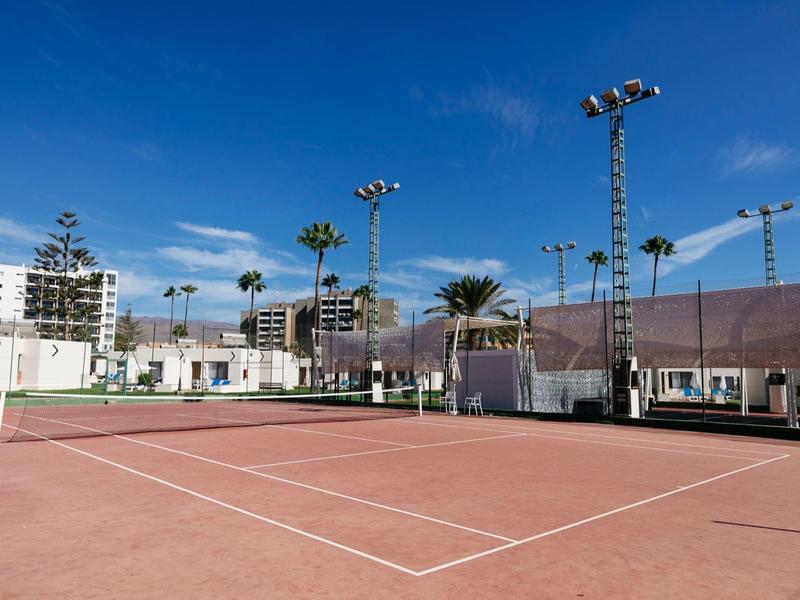 Outdoor tennis court with red clay surface and surrounding urban scenery under blue sky.