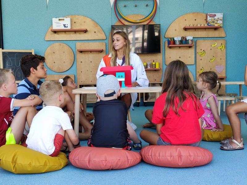 Children sit on colorful cushions in a circle with a caregiver in a bright room.