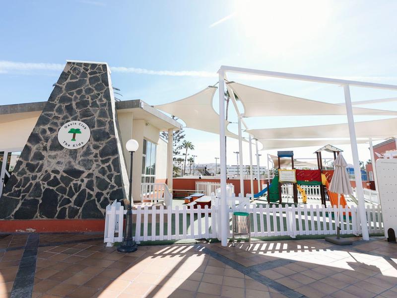 Hotel outdoor area with stone oven, white fence, and playground under sunshades.