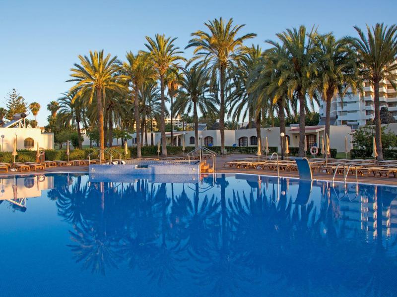 Large pool with palm trees and lounge chairs in front of a hotel at sunset.