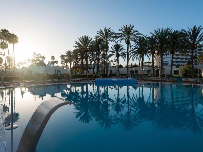 Pool with clear reflective water, surrounded by palm trees and buildings at sunset.