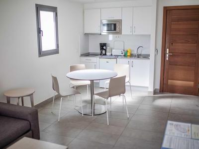 Modern lounge area with white kitchenette, round dining table, and neutral-colored chairs.