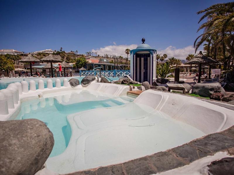Large pool with slides and lounge chairs at a tropical resort under clear skies.