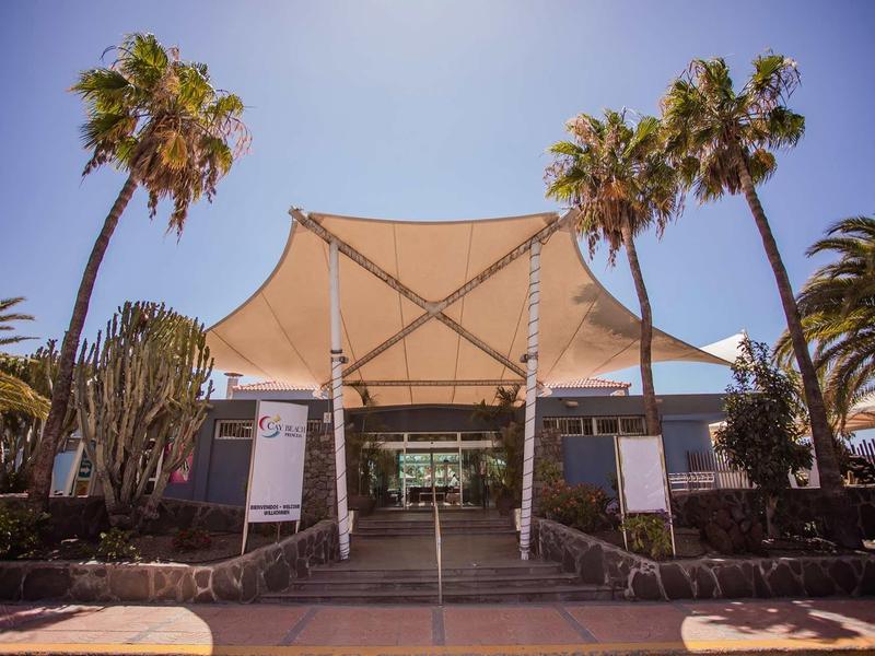 Hotel entrance with a large sunshade canopy and palm trees surrounding it.