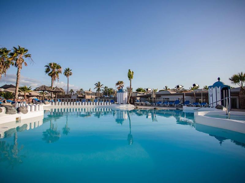 Large outdoor pool with clear blue water, surrounded by palm trees and lounge chairs under a blue sky.