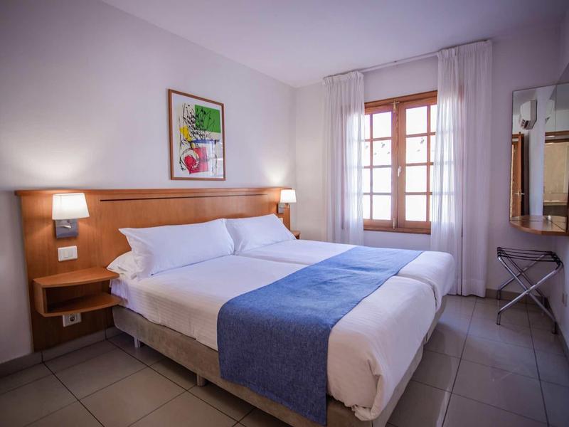 Bright hotel room with double bed, blue bed runner, and window with white curtains.