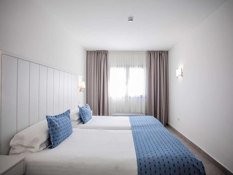 Simple hotel room with two single beds, blue pillows, and white walls.