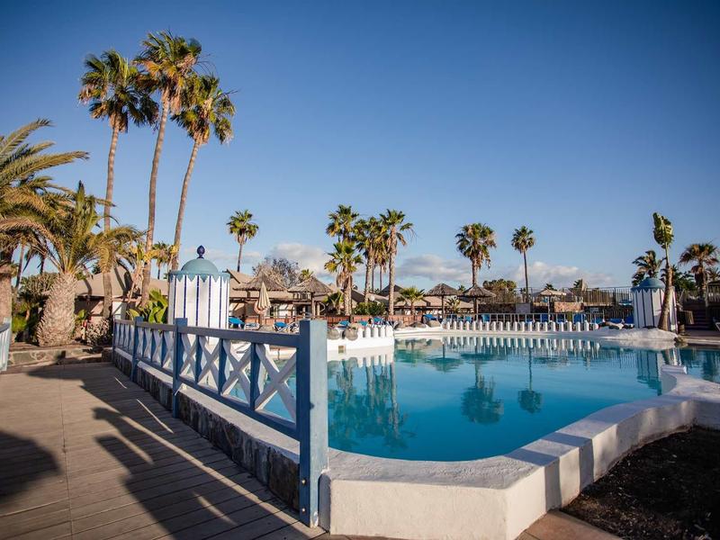 Hotel pool with palm trees and sun umbrellas under a clear blue sky.