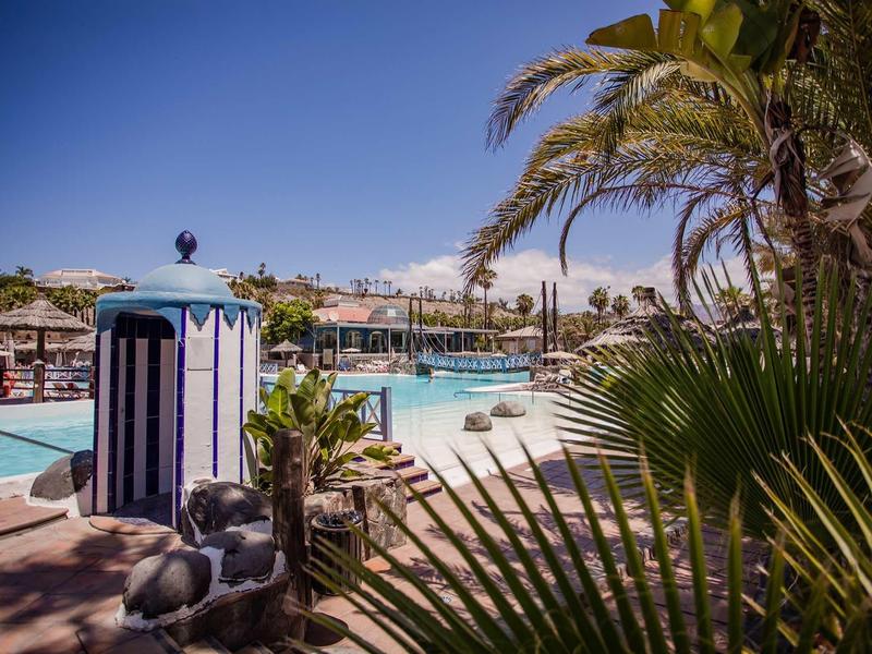 Hotel pool with palm trees and sun umbrellas under a clear blue sky.