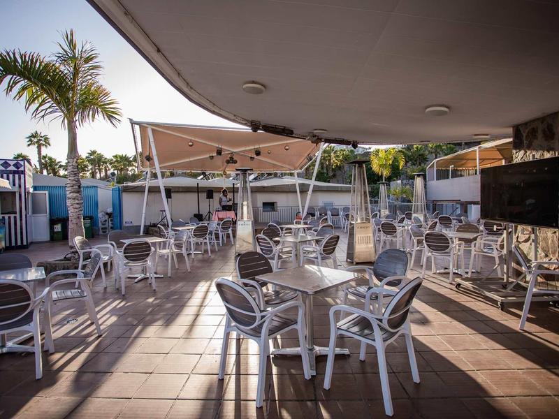 Outdoor terrace with white chairs and tables under sunshades in a tropical setting.