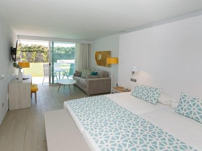 Chambre d'hôtel lumineuse avec lit double, canapé, bureau et vue sur balcon verdoyant.