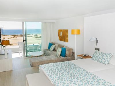 Chambre d'hôtel moderne avec grand lit, canapé, vue sur mer depuis une terrasse vitrée.