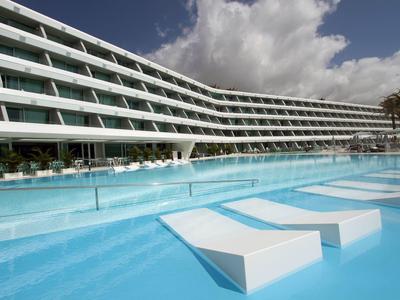 Piscine bleue avec des chaises longues intégrées devant un bâtiment d'hôtel moderne en courbe.