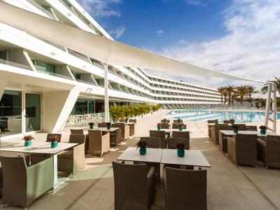 Terrasse d'un hôtel moderne avec tables, chaises et piscine sous un ciel partiellement nuageux.