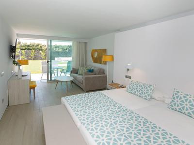 Chambre d'hôtel lumineuse avec lit double, canapé et balcon donnant sur un paysage verdoyant.