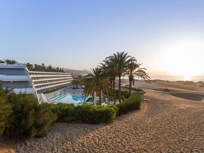 Modern hotel with pool and palm trees next to sandy dunes at sunset.