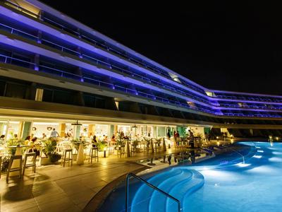Piscine bleue éclairée la nuit devant un hôtel avec terrasse animée.